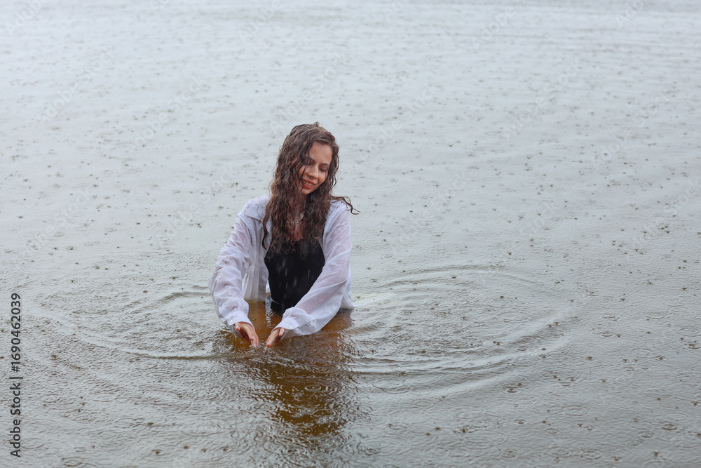 Fototapeta premium Beautiful young brunette woman in black swimsuit and white shirt standing in water of the sea near sandy shore in rainy weather