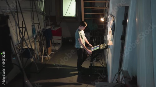 Wallpaper Mural Man in casual clothes stands in dim industrial workspace operating touchscreen control panel under warm desk lamp beside bulletin board filled with notes, while cables and ladders surround the scene Torontodigital.ca