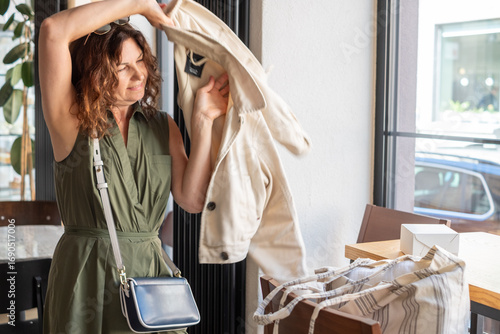 Mid-aged woman with curly hair wearing a stylish green dress is joyfully putting on a light jacket in an urban cafe, capturing the essence of outdoor fashion and lifestyle