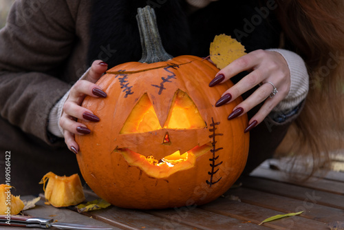 Close-up view of woman with red long sharp fingernails touching orange carved Halloween pumpkin with glowing face lying on wooden table in autumn park. Handheld video. Holiday's symbol theme.