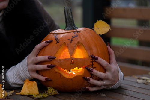 Close-up view of woman with red long sharp fingernails touching orange carved Halloween pumpkin with glowing face lying on wooden table in autumn park. Handheld video. Holiday's symbol theme.