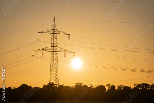 Transmission tower or high-voltage electricity pylon at bright sunset or sunrise, landscape format, symbol of energy and power generation in Germany.