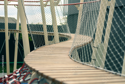Curved wooden bridge with safety netting in a playground, captured in perspective. Outdoor adventure structure designed for children’s play and climbing activities.