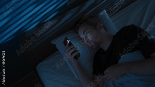 Tired young man lies in bed at night, staring at his smartphone screen, doom scrolling through social media, unable to sleep struggling with sleep disorder or distraction by overthinking. Copy space