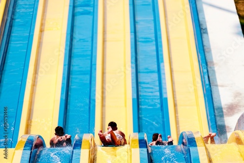 Three young boys are enjoying a sunny day at an Aqua Park. They are racing down the blue and yellow water slides, splashing in the water as they descend