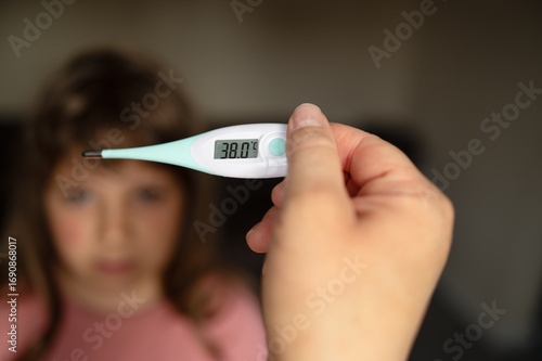 Digital thermometer displaying high temperature, in front of a blurred girl in the background, Concept: child illness, healthcare, cold and flu.