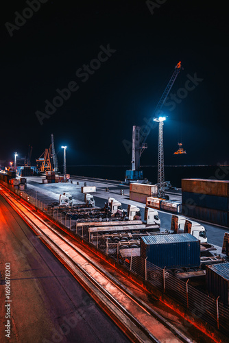 Industrial cargo port at night with shipping containers, cranes, and trucks illuminated by artificial lights, highlighting global trade, logistics, and heavy transportation industry