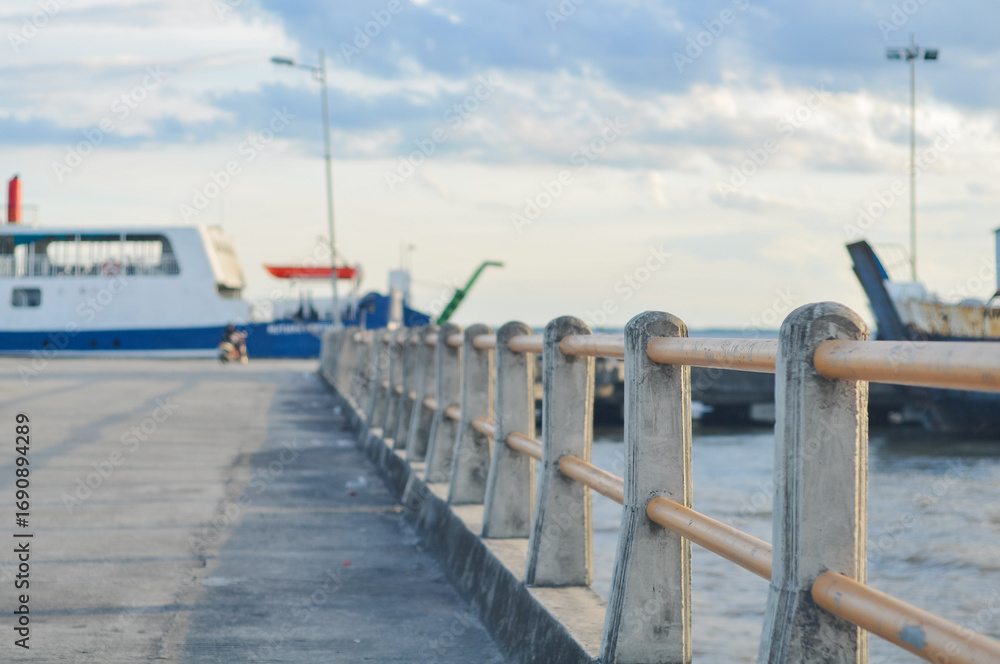 Naklejka premium Concrete pier with safety railing at a seaport. Blurry view of a ferry ship and sea water in the background under cloudy sky. Transportation, travel, and maritime industry concept.