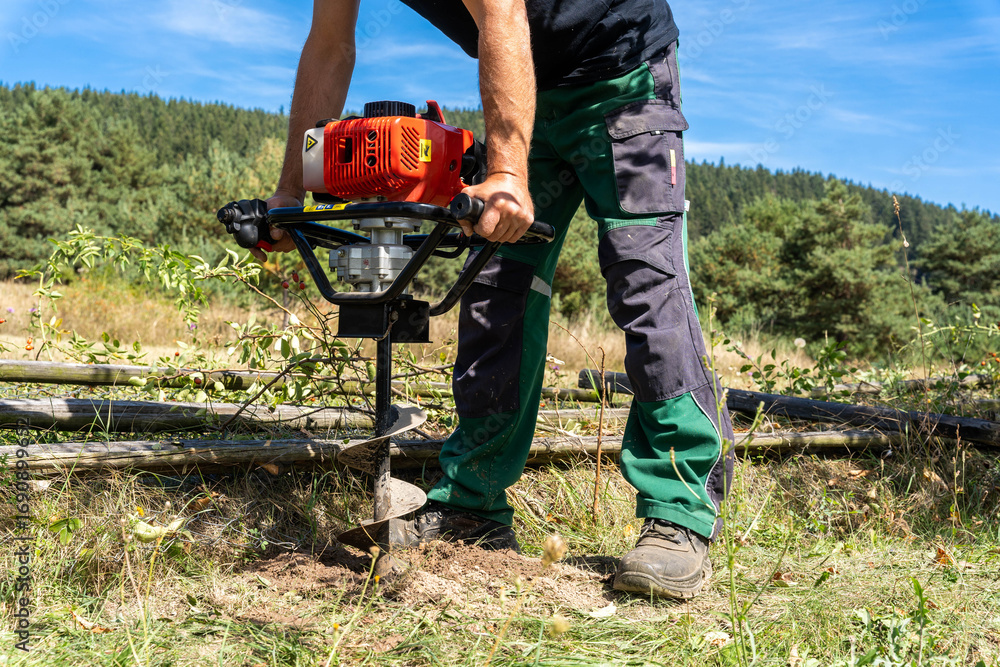 Obraz premium Worker drilling hole for wooden post with handheld gasoline powered auger drill in very rough, dry soil.