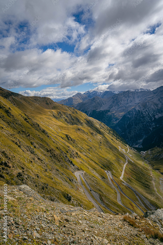 Fototapeta premium Stelvio Pass serpentine road weaving up alpine mountainside with panoramic view of peaks and valleys under shifting clouds dramatic scenery