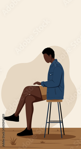 Young Black man in casual attire, focused on his laptop while sitting on a stool. Represents remote work, freelance lifestyle, productivity, or online education.