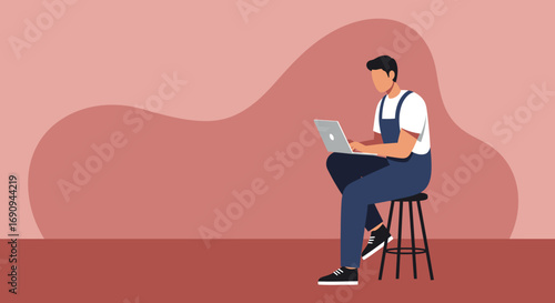 Young man in overalls working intently on a laptop while seated on a stool. Concept of remote work, online learning, or modern tech use. Copy space.