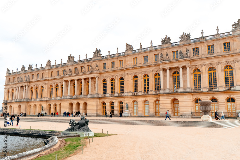 Fototapeta premium Visitors explore the grand exterior of Palatul Versailles, surrounded by manicured gardens and elegant sculptures, under a cloudy sky in France's historic site.