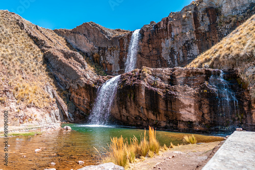 Tourist view of a two-level waterfall in Marcapomacocha, Peru, cascading into a clear emerald pool surrounded by cliffs and Andean grasslands.