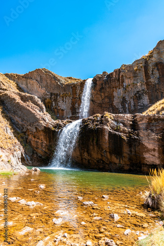 Tourist view of a two-level waterfall in Marcapomacocha, Peru, cascading into a clear emerald pool surrounded by cliffs and Andean grasslands.