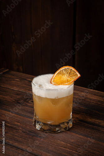 Citrus yellow cocktail with airy foam and dried orange garnish served in short glass on rustic wooden table