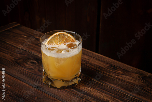 Classic whiskey sour cocktail with ice cubes and dried orange slice served in lowball glass on rustic wooden table