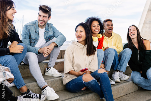 Multiracial group of young teenage student friends laughing together sitting outdoor. Youth community concept