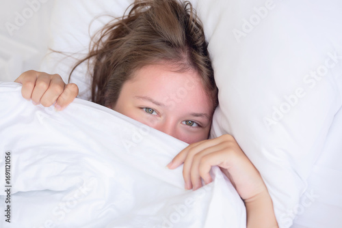 Young funny woman hiding her face under the bed blanket at home