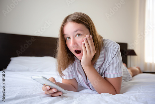 An attractive cheerful young woman is lying on a bed at home, holding a mobile phone in her hands and talking. Surprised expression on his face