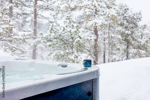 Contrasts of heat and frost — a steaming outdoor jacuzzi with a ceramic mug invites warmth and relaxation amid a serene snow-covered forest. Winter indulgence meets Nordic nature.