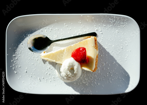 A slice of cheesecake with a dollop of whipped cream and a strawberry and spoon on a white plate dusted with powdered sugar against a black background
