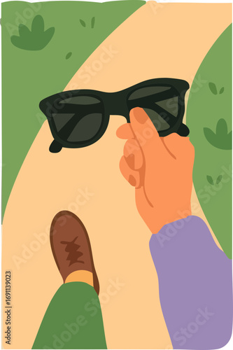 person walking on a path holding black sunglasses, with one brown shoe and green pants visible.