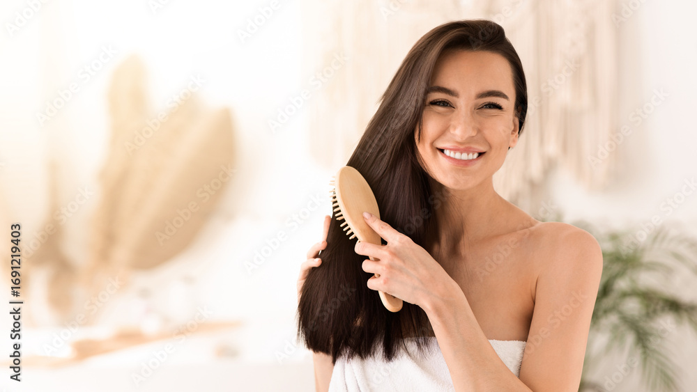 Naklejka premium Woman makes hair care in home, treatment and spa day at leisure time. Happy young cute brunette female wrapped in towel, combing long healthy hair with comb, in light bathroom interior in morning