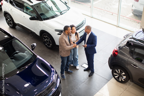 Happy African American couple shaking hands with car dealer in modern showroom, celebrating successful purchase, lifestyle and finance moment of auto shopping