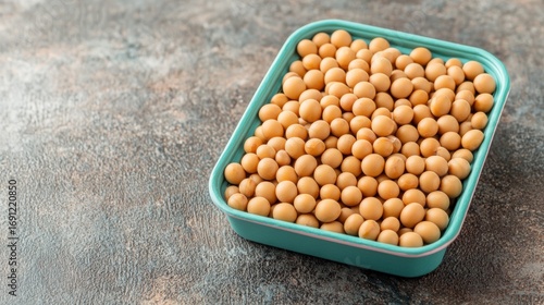 A teal container filled with small, round beige soybeans placed on a textured gray surface.
