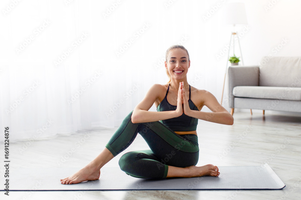Fototapeta premium Practice yoga in morning, stretching, body care at home, breathing exercises during covid-19 quarantine. Glad young blonde female in sportswear work out, in minimalist living room interior, copy space