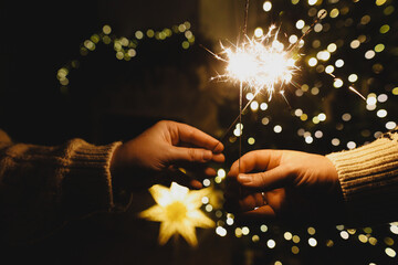 Hands holding fireworks against christmas tree lights bokeh in evening cozy living room. Happy New Year eve! Atmospheric family celebration with burning sparklers in hands close up