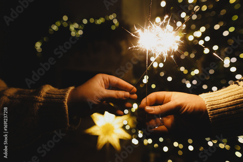 Hands holding fireworks against christmas tree lights bokeh in evening cozy living room. Happy New Year eve! Atmospheric family celebration with burning sparklers in hands close up