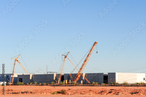 Cranes position steel and concrete panels during construction of a large industrial building, with heavy equipment and structural framework visible at an active worksite under clear sky conditions