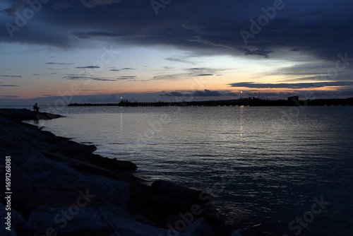 Calm sea at sunset. Dark clouds in the sky. A lone fisherman sits on a rocky shore. An illuminated pier in the background.