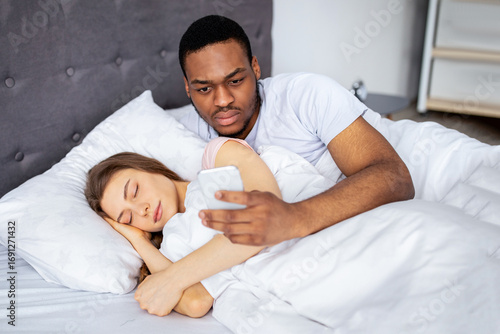 Fotografie Jealous black husband checking his wife's smartphone, reading correspondence while she is asleep in bed at home
