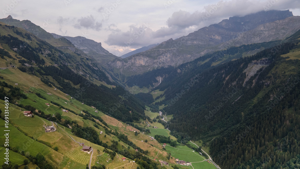 Obraz premium Swiss Alps with houses, winding road, clouds, and overcast sky, captured by drone. Schweizer Alpen mit Häusern, kurviger Straße, Wolken und bedecktem Himmel, aufgenommen mit Drohne.