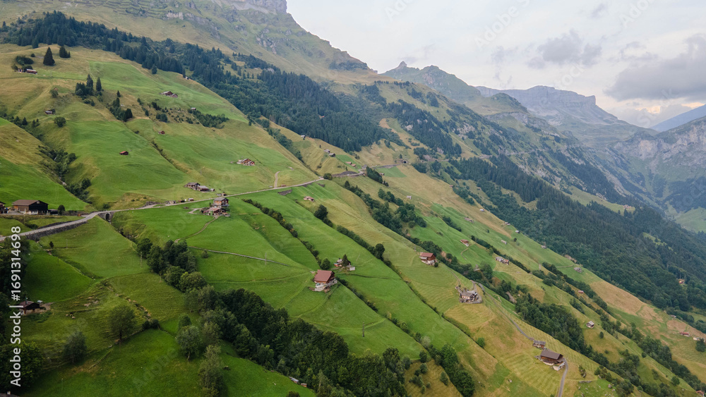 Obraz premium Swiss Alps with houses, winding road, clouds, and overcast sky, captured by drone. Schweizer Alpen mit Häusern, kurviger Straße, Wolken und bedecktem Himmel, aufgenommen mit Drohne.