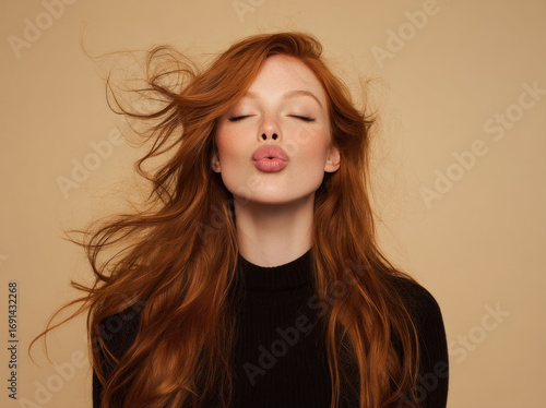 Portrait of a young woman with long red hair blowing in the wind, lips puckered in a playful kiss, wearing a dark sweater against a warm beige backdrop.