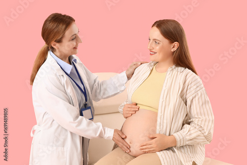 Young pregnant woman with doctor on couch against pink background