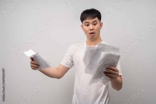 A stressed and anxious young man expresses shock and despair while reviewing numerous overdue bills and financial statements. Concept for business, finance, advertising, and lifestyle topics.