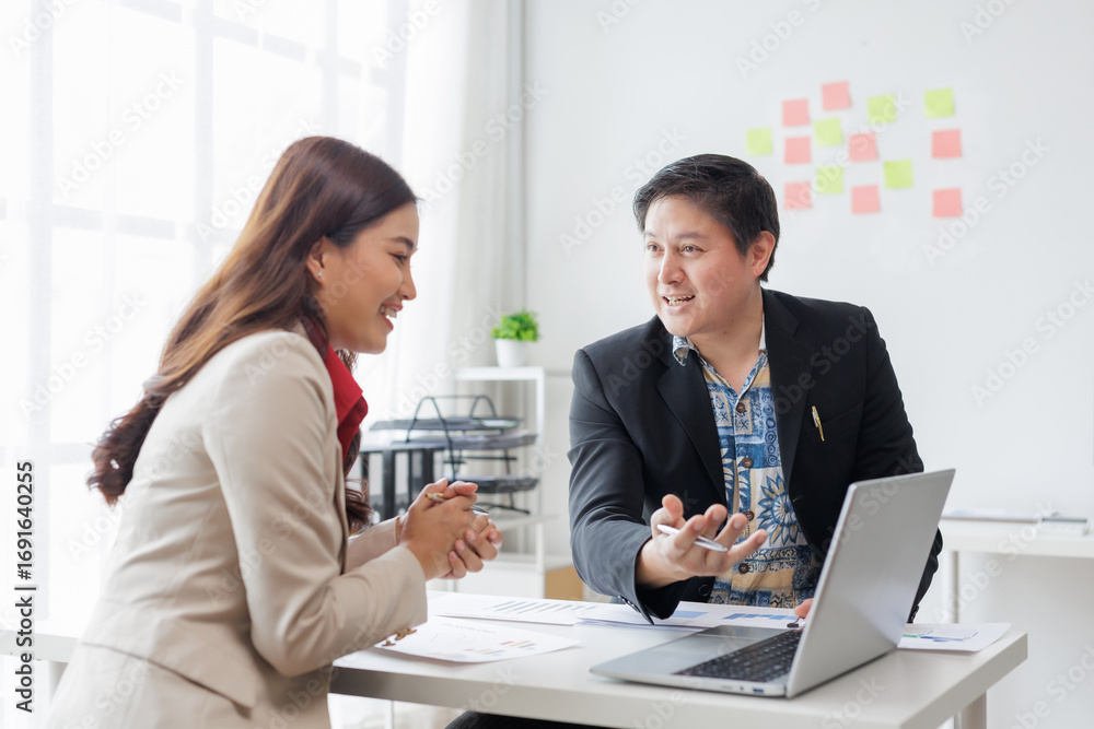 Fototapeta premium Asian businesspeople discussing during meeting in modern office using laptop and documents, analyzing financial charts, collaborating and sharing ideas