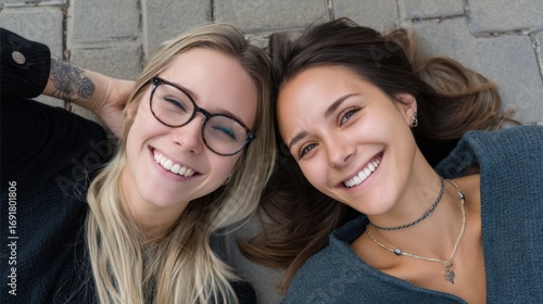 Overhead shot of joyful sisters, one wearing glasses, the other with a tattoo, lying on footpath 