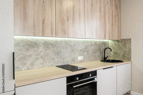 modern kitchen features light wood cabinets, a marble backsplash illuminated by strip lighting, black fixtures, and built-in appliances