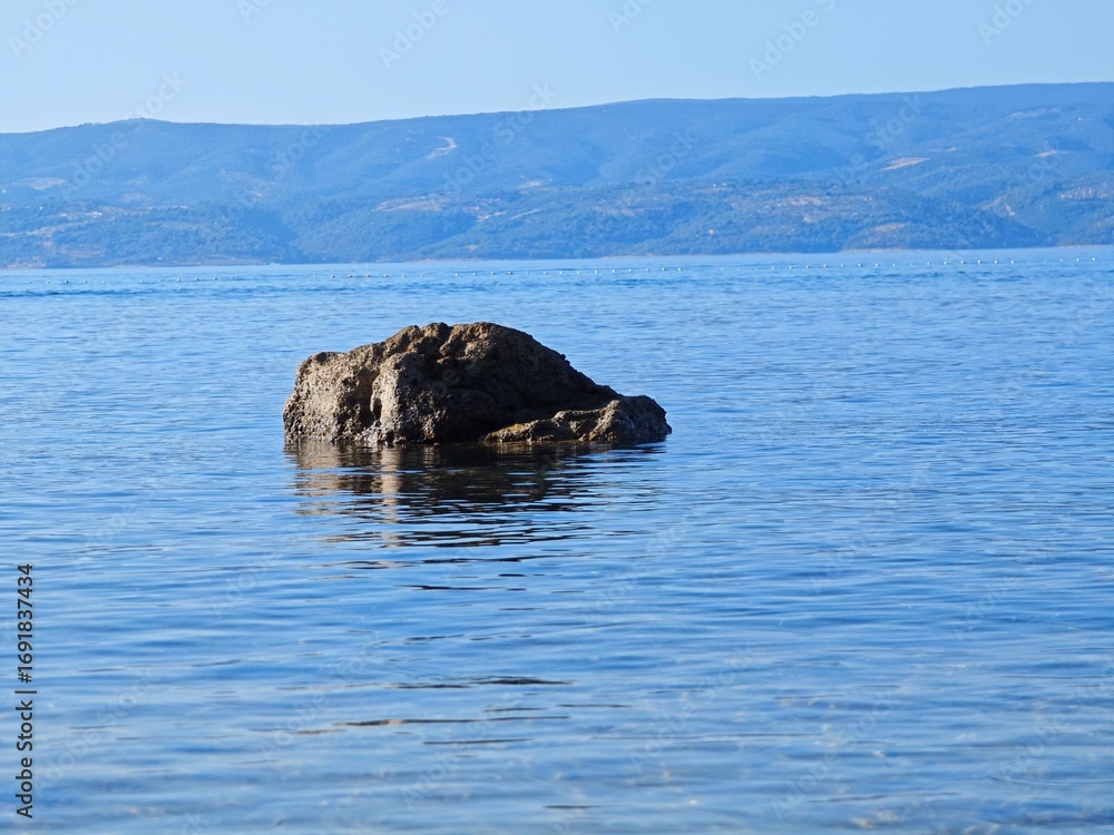 Fototapeta premium Calm waters reflect a solitary rock near the shoreline under a clear blue sky in summertime