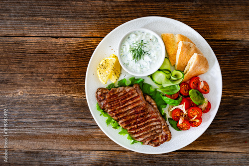 Grilled beef steak with tzatziki sauce, pita bread and fresh vegetables on wooden table	
