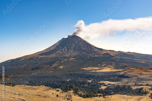 A mountain with a volcano on top and a cloud of smoke rising from it. The sky is clear and blue. Iztacihuatl popocatepetl volcano mexico,