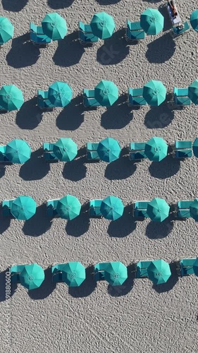 Vertical 4K top-down aerial video of turquoise umbrellas and lounge chairs arranged in perfect rows on Miami Beach, Florida, with morning sun casting long shadows, creating a vibrant geometric beach s
