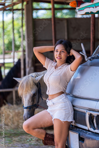 A beautiful young woman is standing in front of a six-wheeled truck on a horse farm.