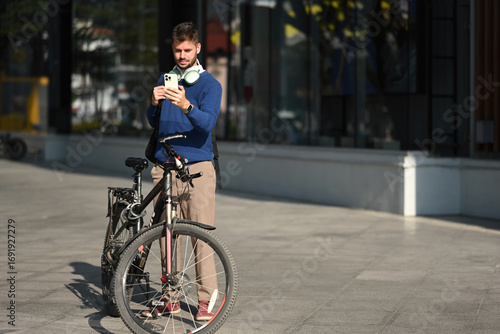 A Professional Caucasian man with backpack and bicycle stands outside, looking at his smartphone, blending fitness, technology, and urban life